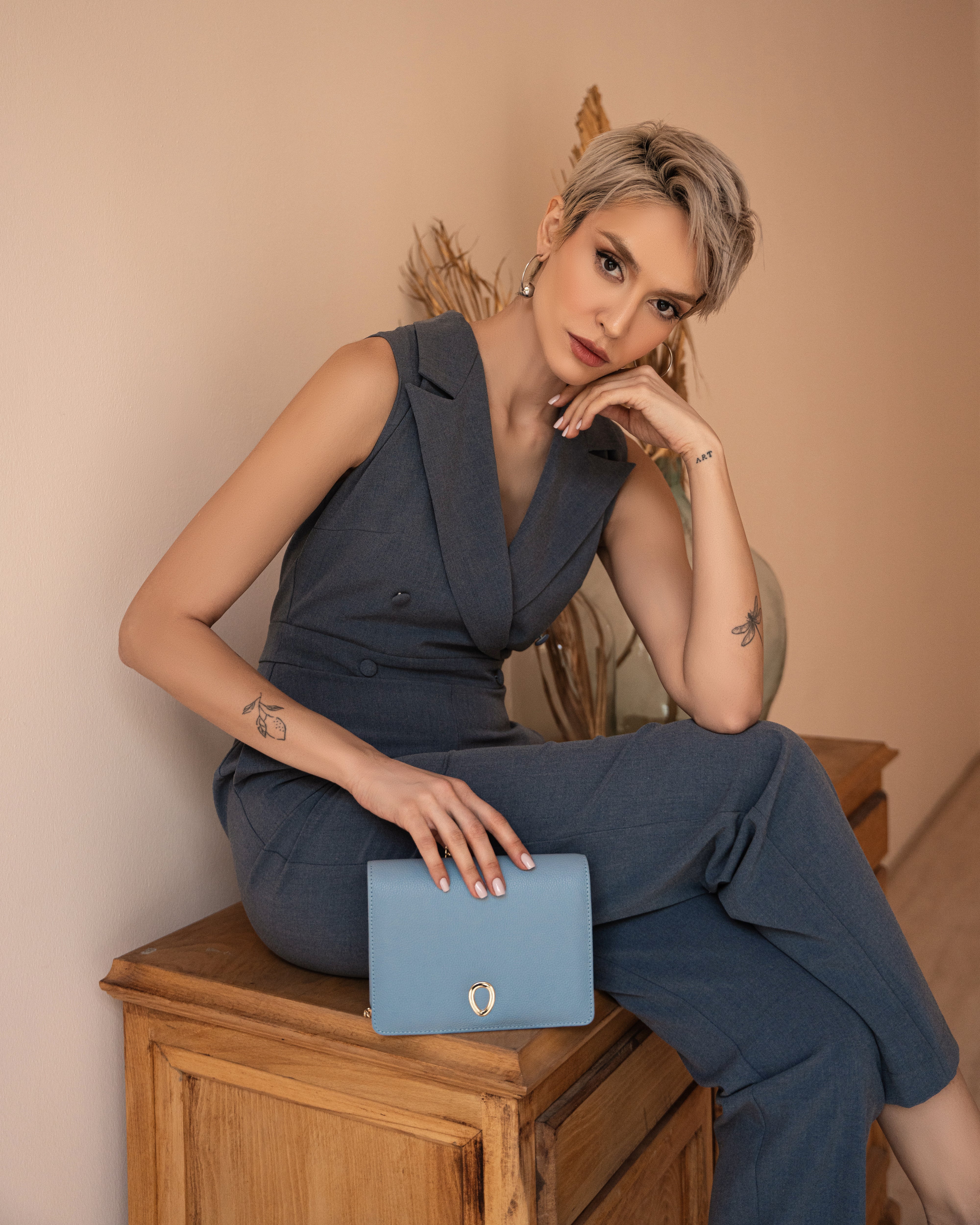 A woman with short blonde hair, dressed in a sleeveless blue pantsuit, sits on a wooden cabinet holding the Vera in leather - Mist Blue evening bag in her lap. Dried plants decorate the background.