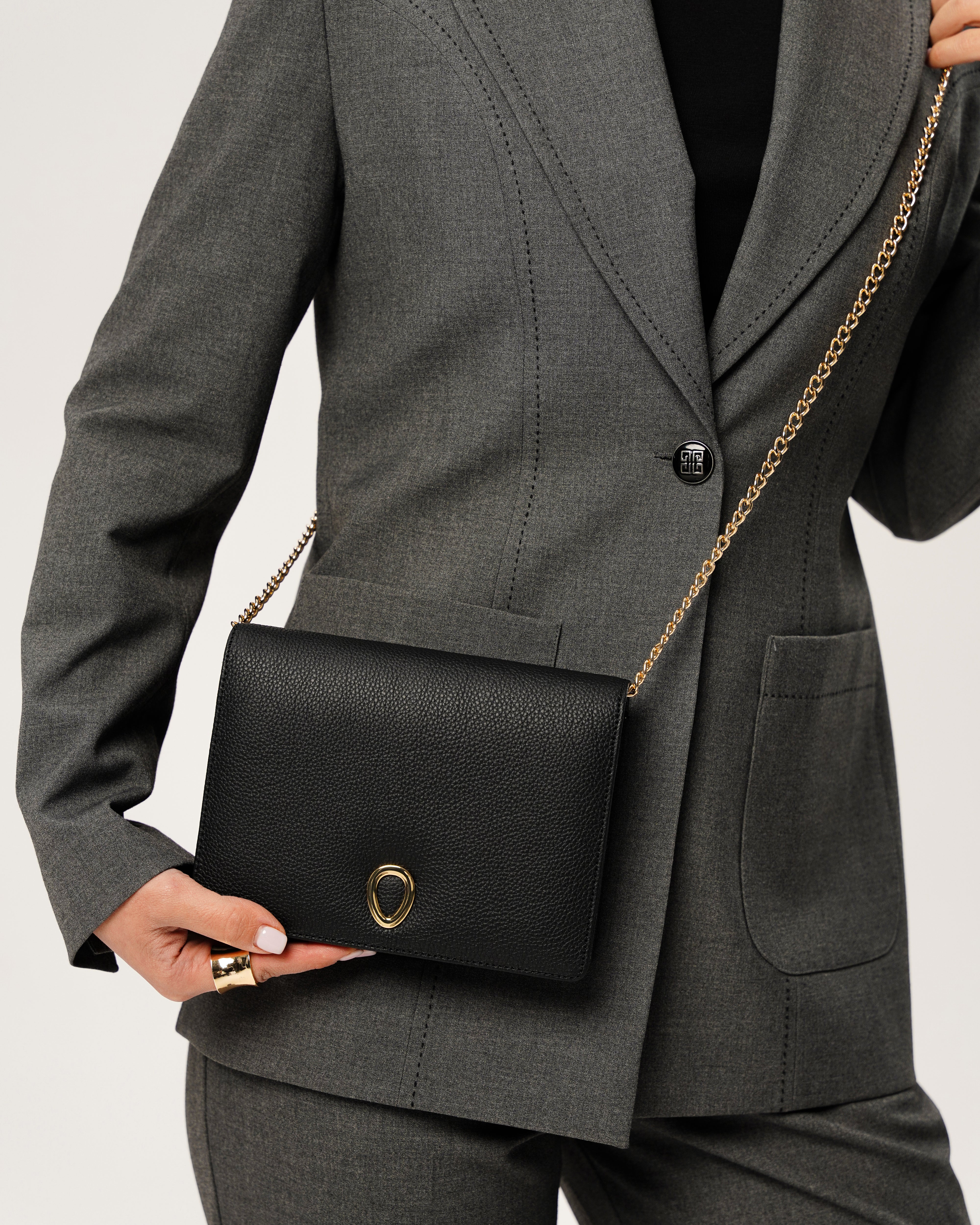 A person in a gray suit holds the Vera in leather - Stone Taupe women’s clutch featuring a gold chain strap and circular gold accent, standing against a light background.