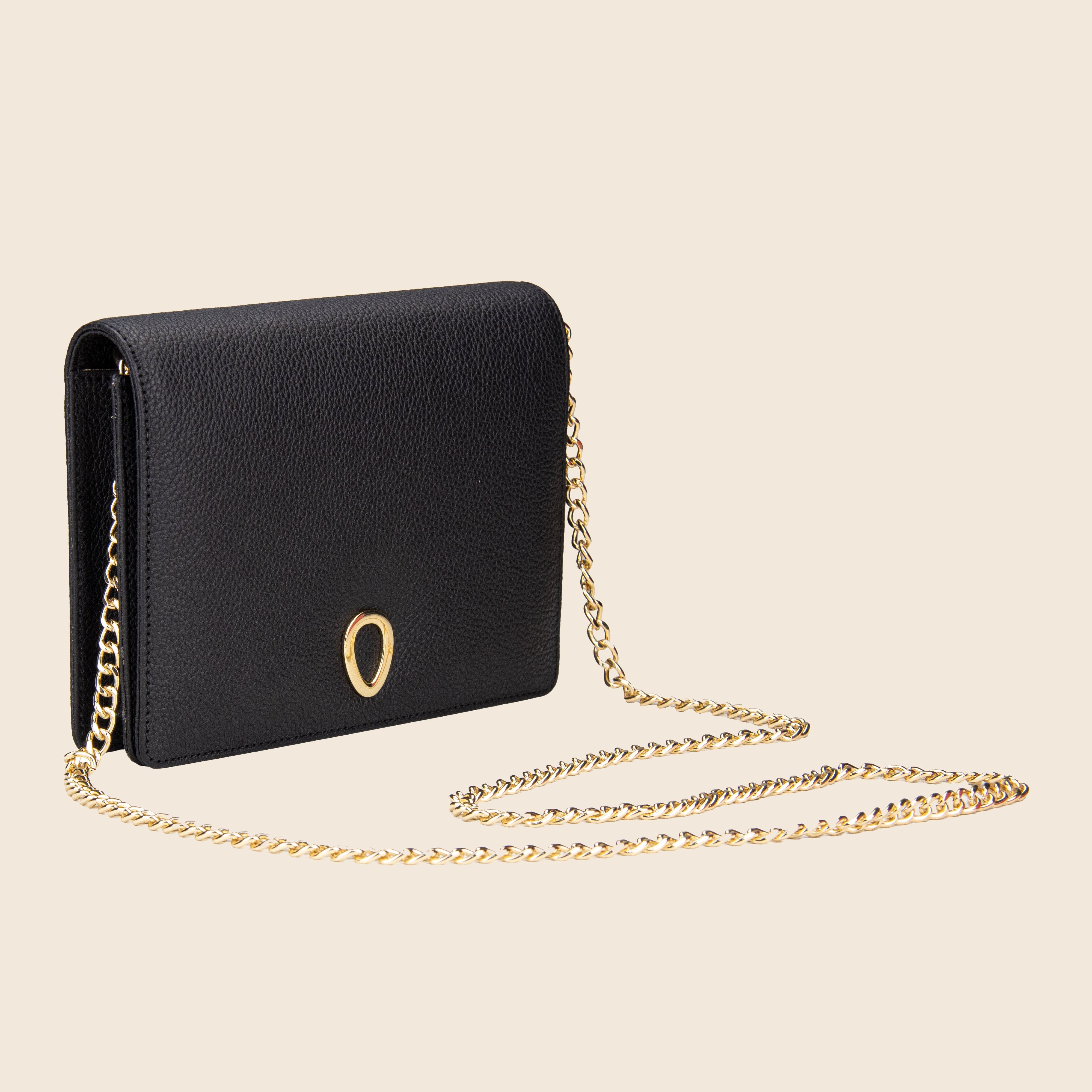The Vera in leather - Classic Black is an elegant women’s bag with a black leather finish, gold chain strap, and a small gold oval accent on the front, shown against a neutral beige background.