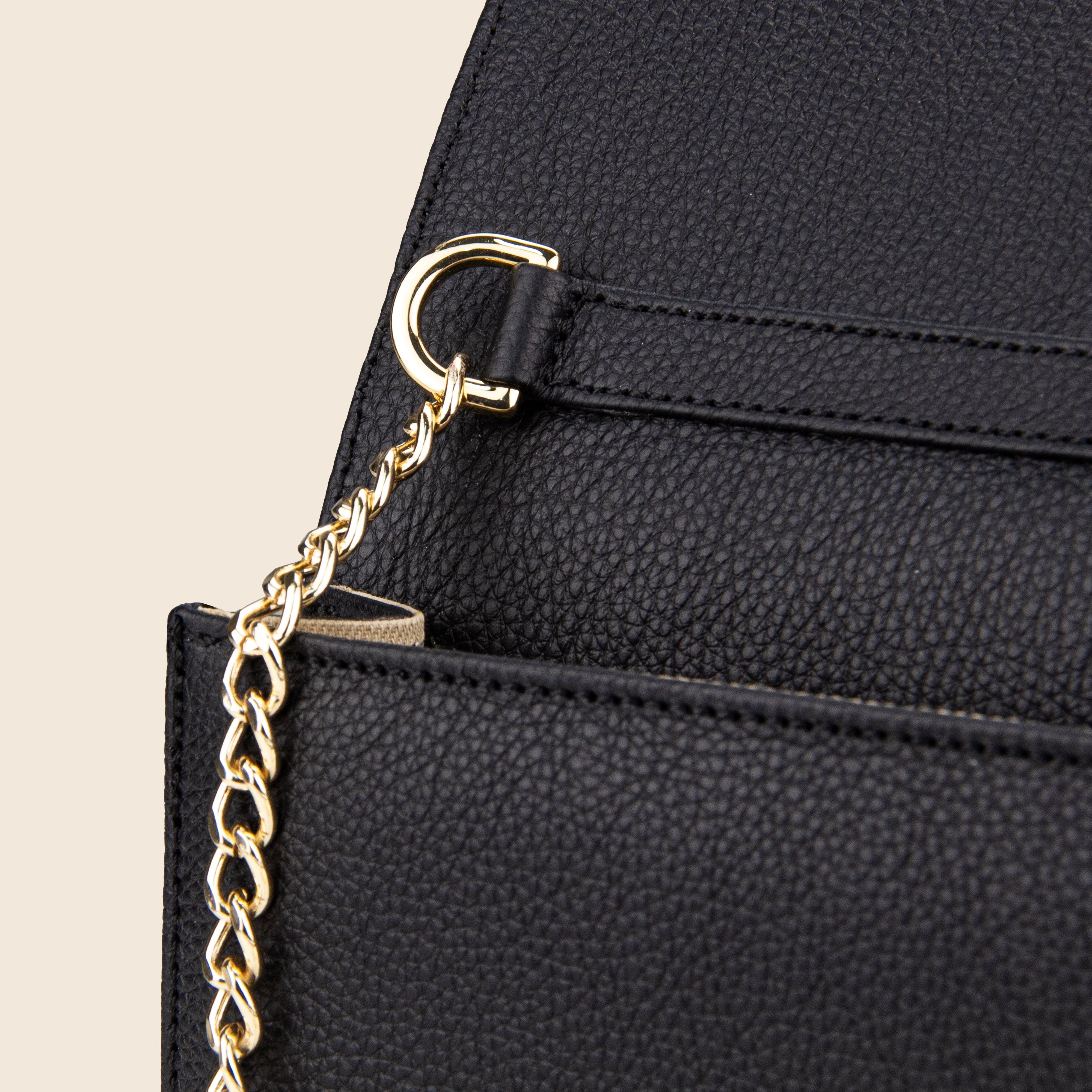 Close-up of the Vera in leather - Classic Black women’s bag, crafted from black textured leather with a gold chain strap attached to a metal ring, set against a beige background.