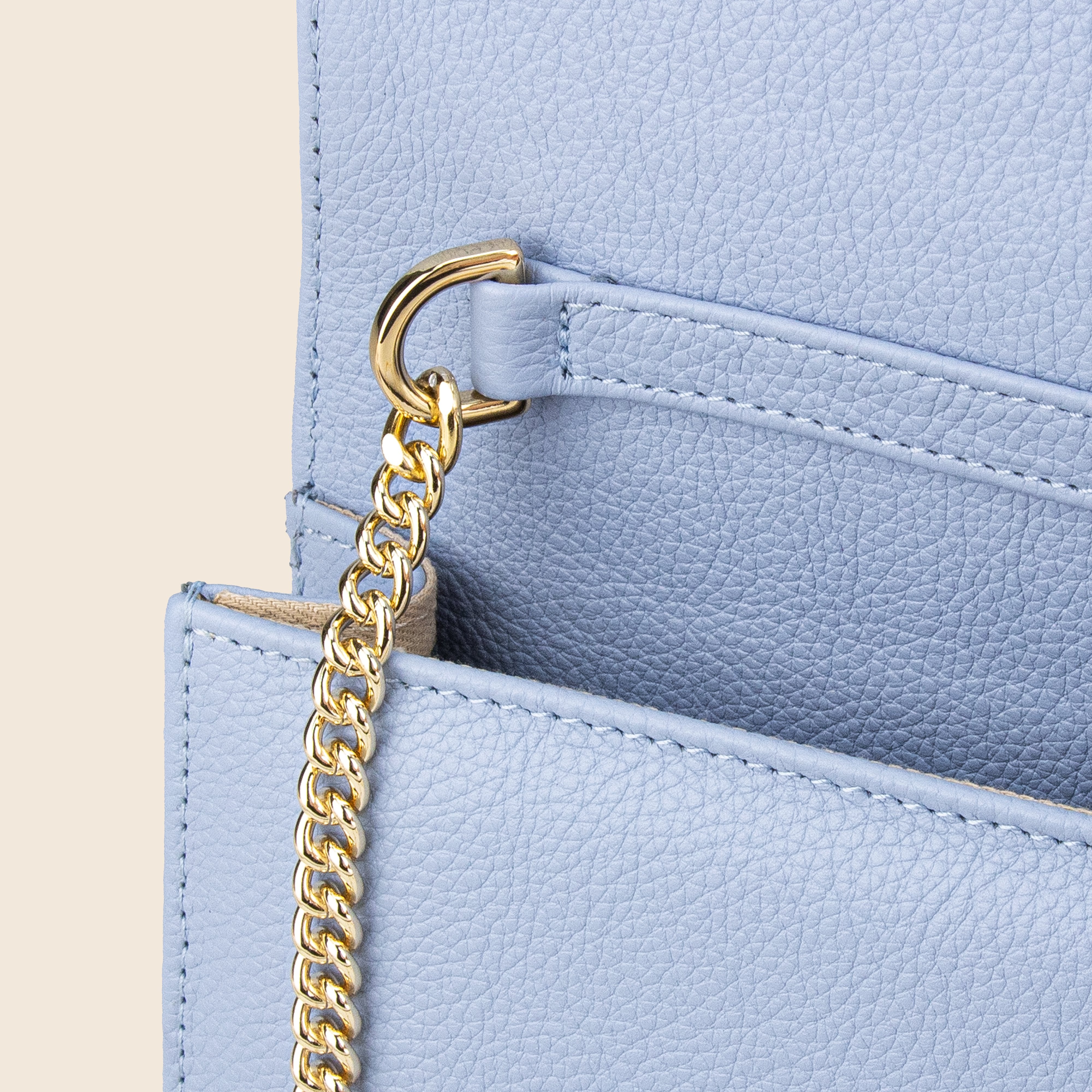 Close-up of Vera in leather - Mist Blue, an elegant evening bag featuring light blue textured leather, a gold chain strap, and flap opening, displayed against a beige background.