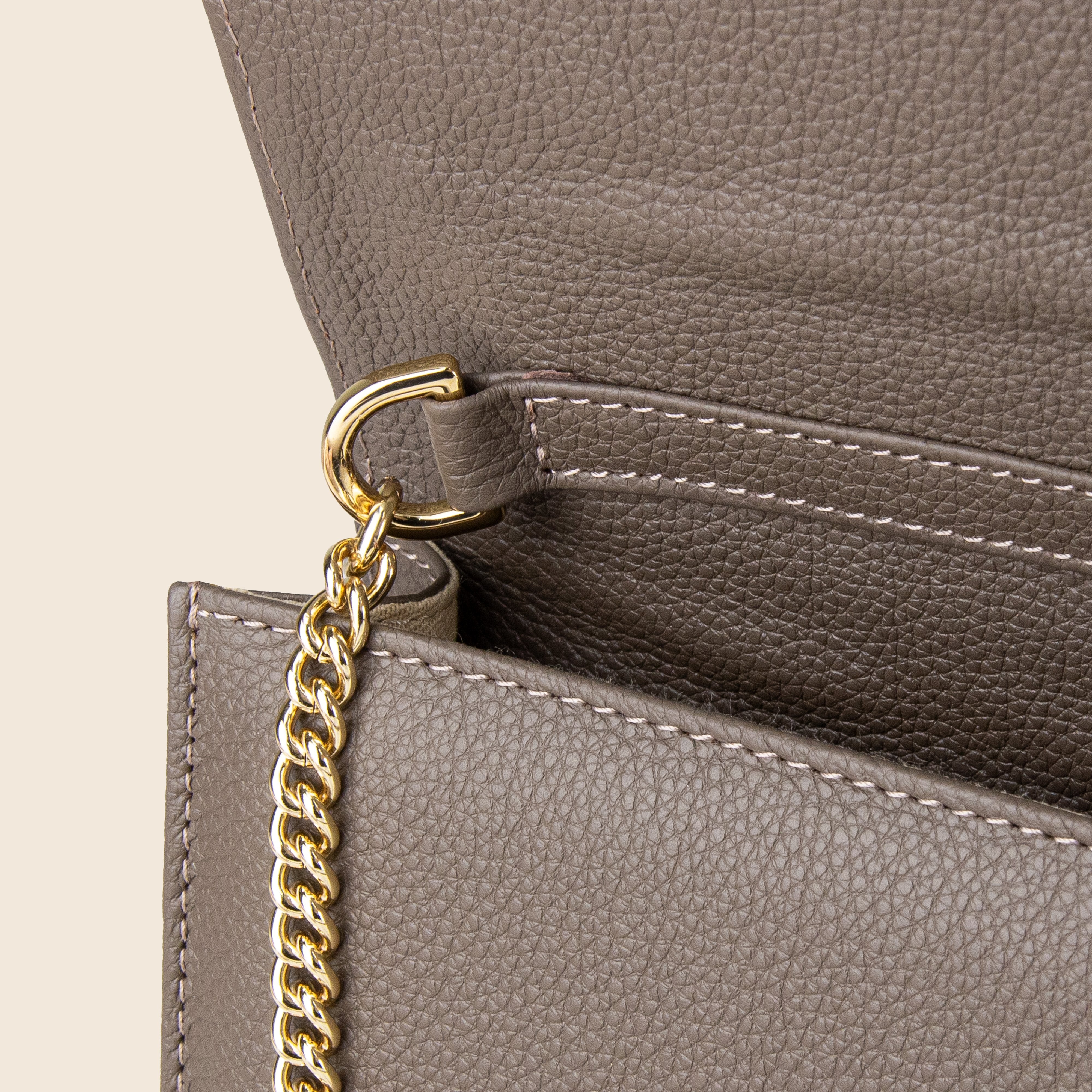 Close-up of the Vera in leather - Stone Taupe clutch, crafted from genuine textured taupe leather. Features a gold chain strap linked to a metal ring beneath the flap, with visible stitching detail along the edge.